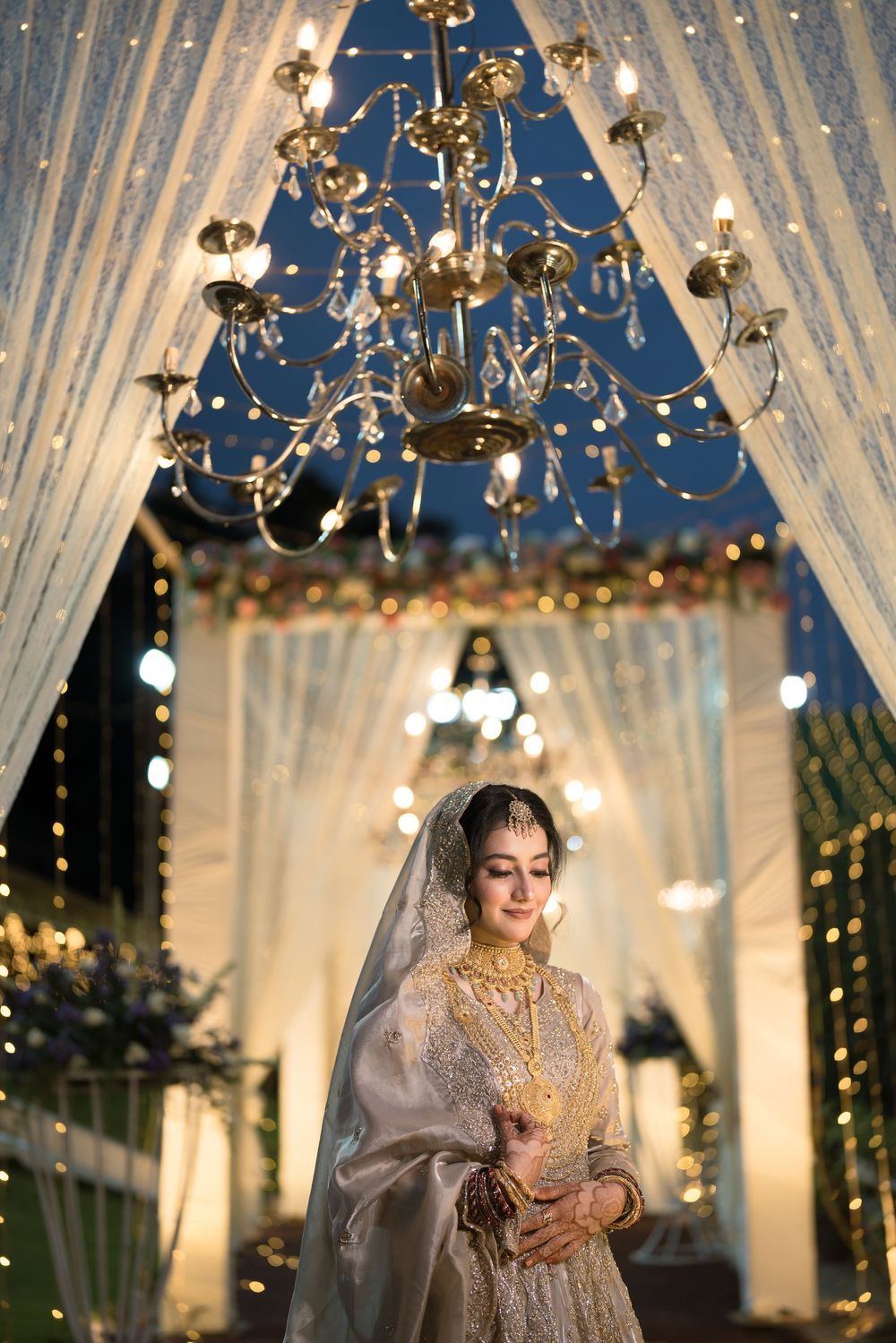 Elegant bride in gold jewelry and dupatta stands under crystal chandelier at decorated wedding venue.