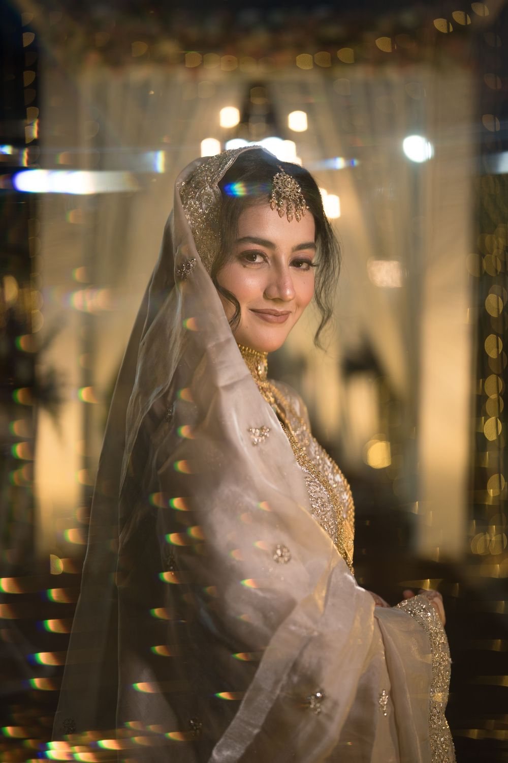 A bride in a white embroidered lehenga and veil with gold jewelry smiles softly amid warm bokeh lights.