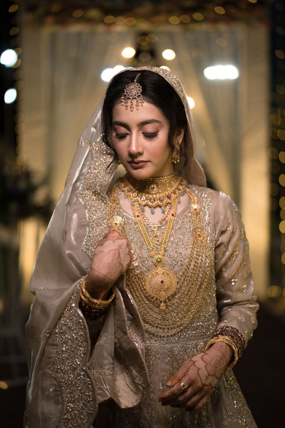 A bride in a gold embroidered lehenga adorned with layered gold jewelry and dupatta, posing gracefully at a wedding ceremony.