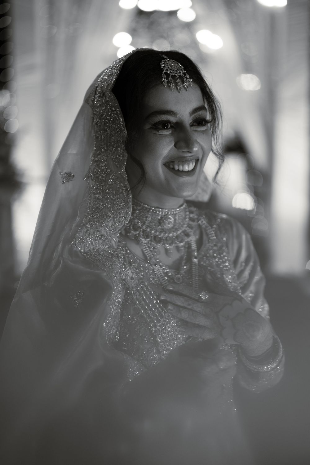 Black and white photo of a smiling Indian bride in embroidered lehenga, veil, and layered necklaces at her wedding.