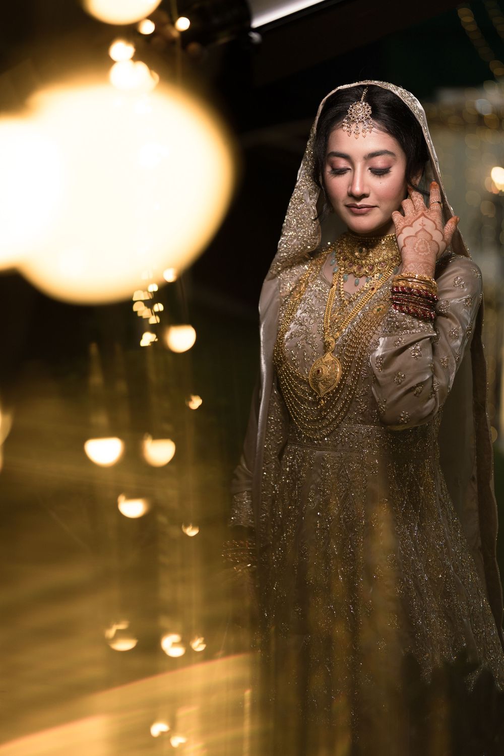 A bride in a gold embroidered lehenga and dupatta adorned with traditional gold jewelry poses gracefully in warm glowing light.