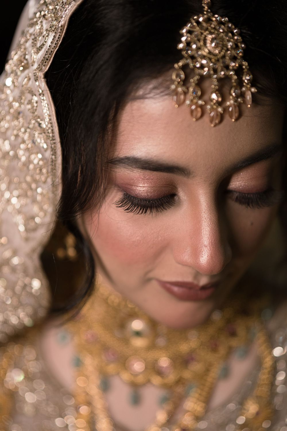 A South Asian bride wearing gold maang tikka, layered kundan jewelry, and embroidered dupatta with elegant bridal makeup.
