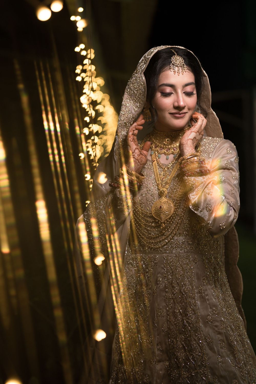 A bride in gold embroidered lehenga and dupatta adorned with traditional jewelry poses gracefully against sparkling lights.