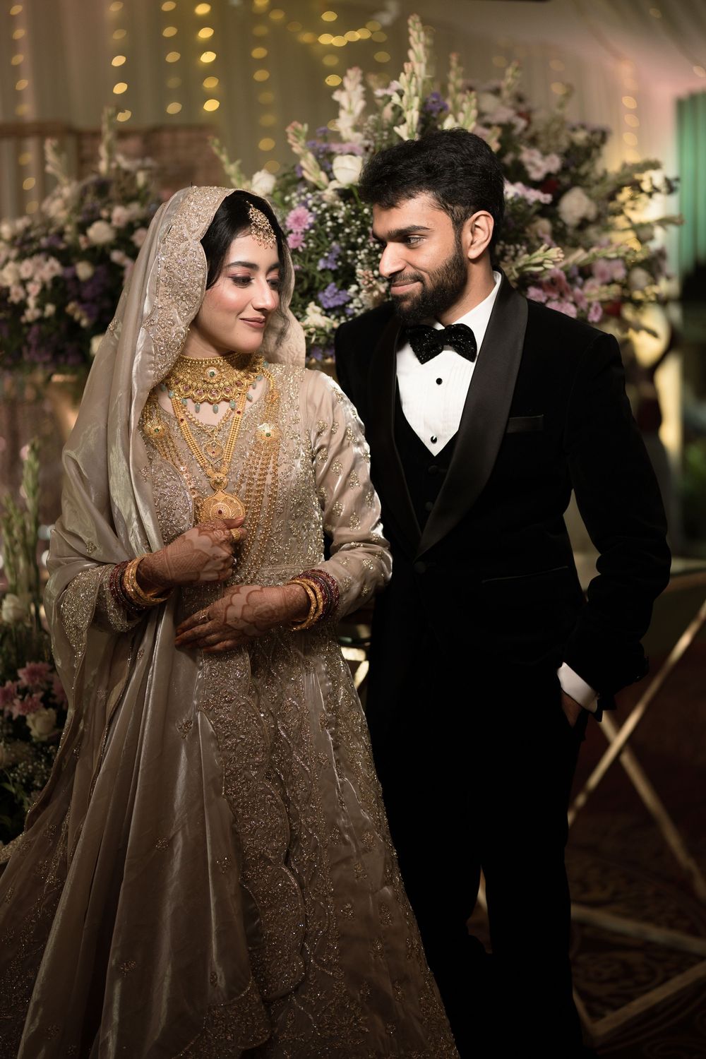 A bride in gold embroidered lehenga with jewelry and groom in black tuxedo pose together at a wedding reception.