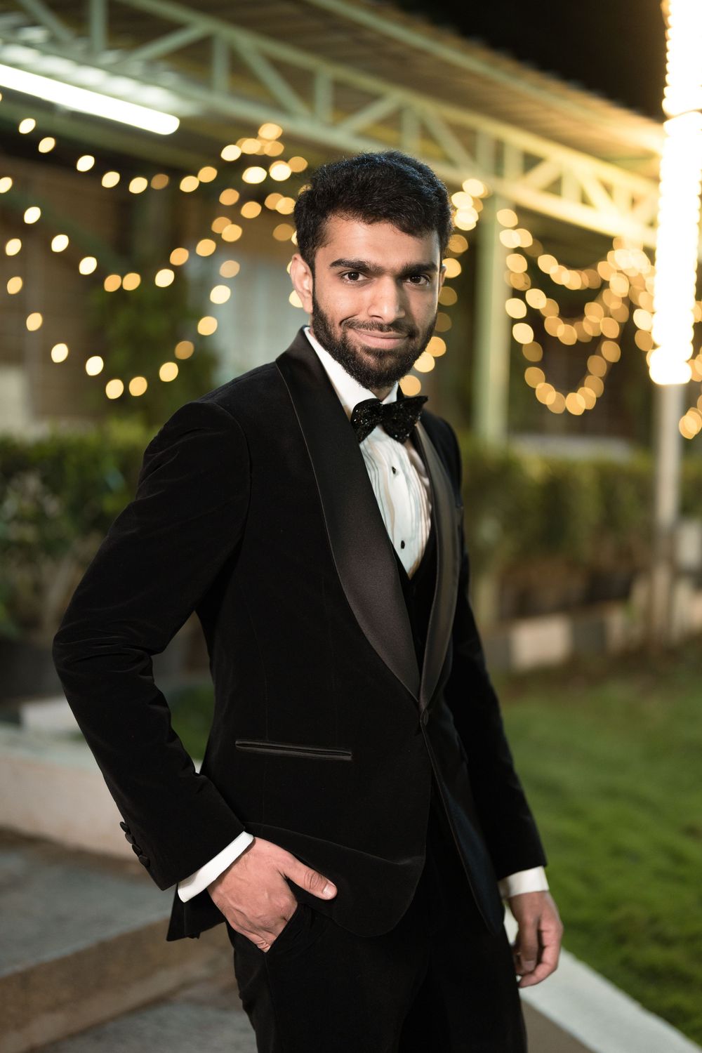 Elegant man in black velvet tuxedo with bow tie posing outdoors at night with bokeh string lights.