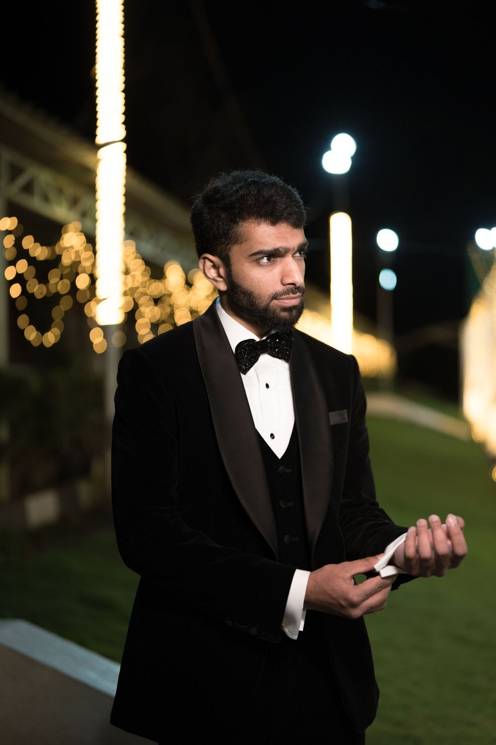 Young man in black tuxedo with bow tie standing outdoors at night with bokeh lights in background.