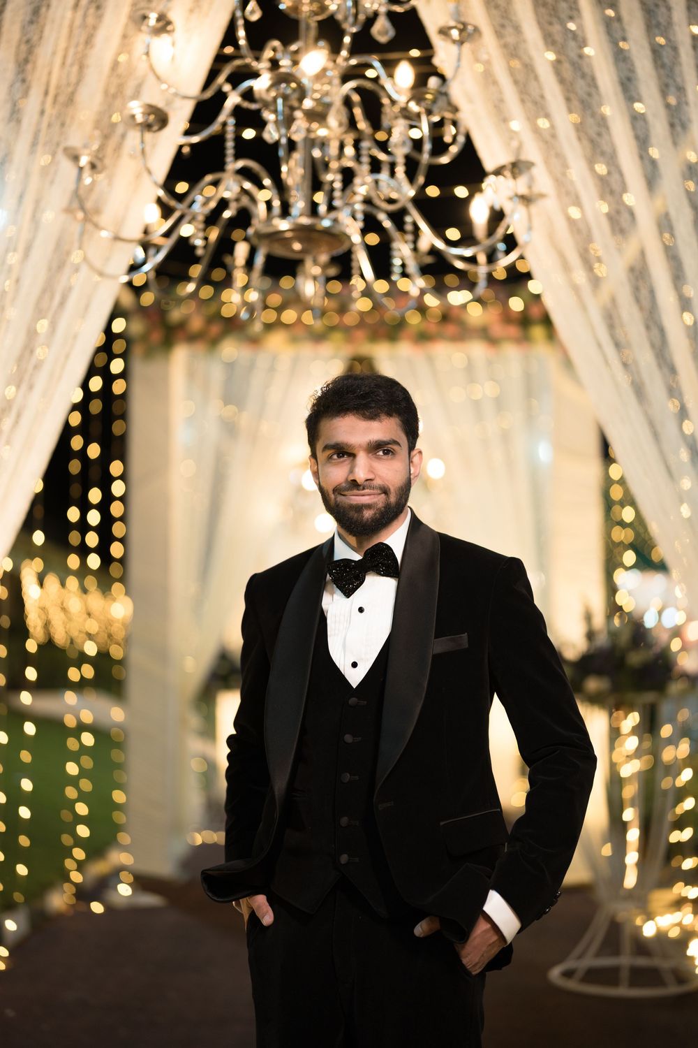Elegant man in black tuxedo with bow tie posing at a glamorous event with chandelier and bokeh lights.