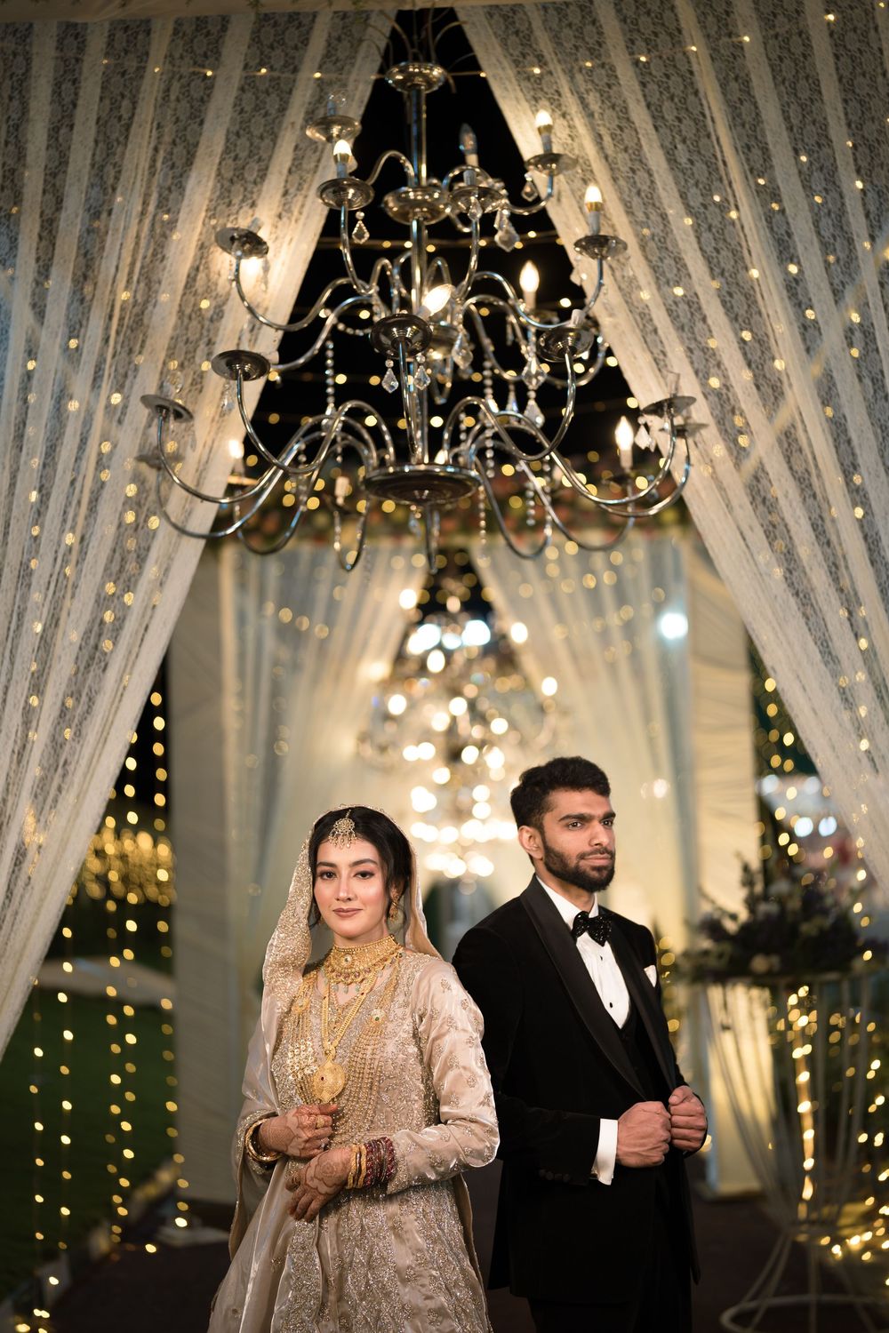 Bride in golden lehenga and groom in black tuxedo pose under chandelier at elegant wedding venue.
