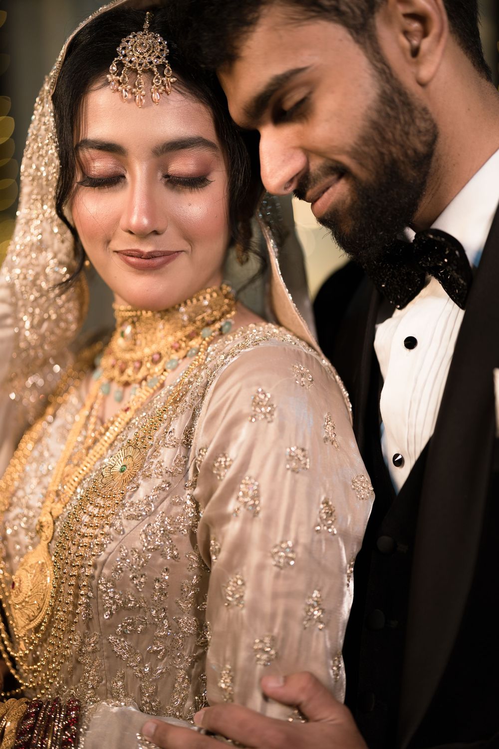 A bride in gold embroidered attire with jewelry and groom in tuxedo share an intimate moment at a South Asian wedding.