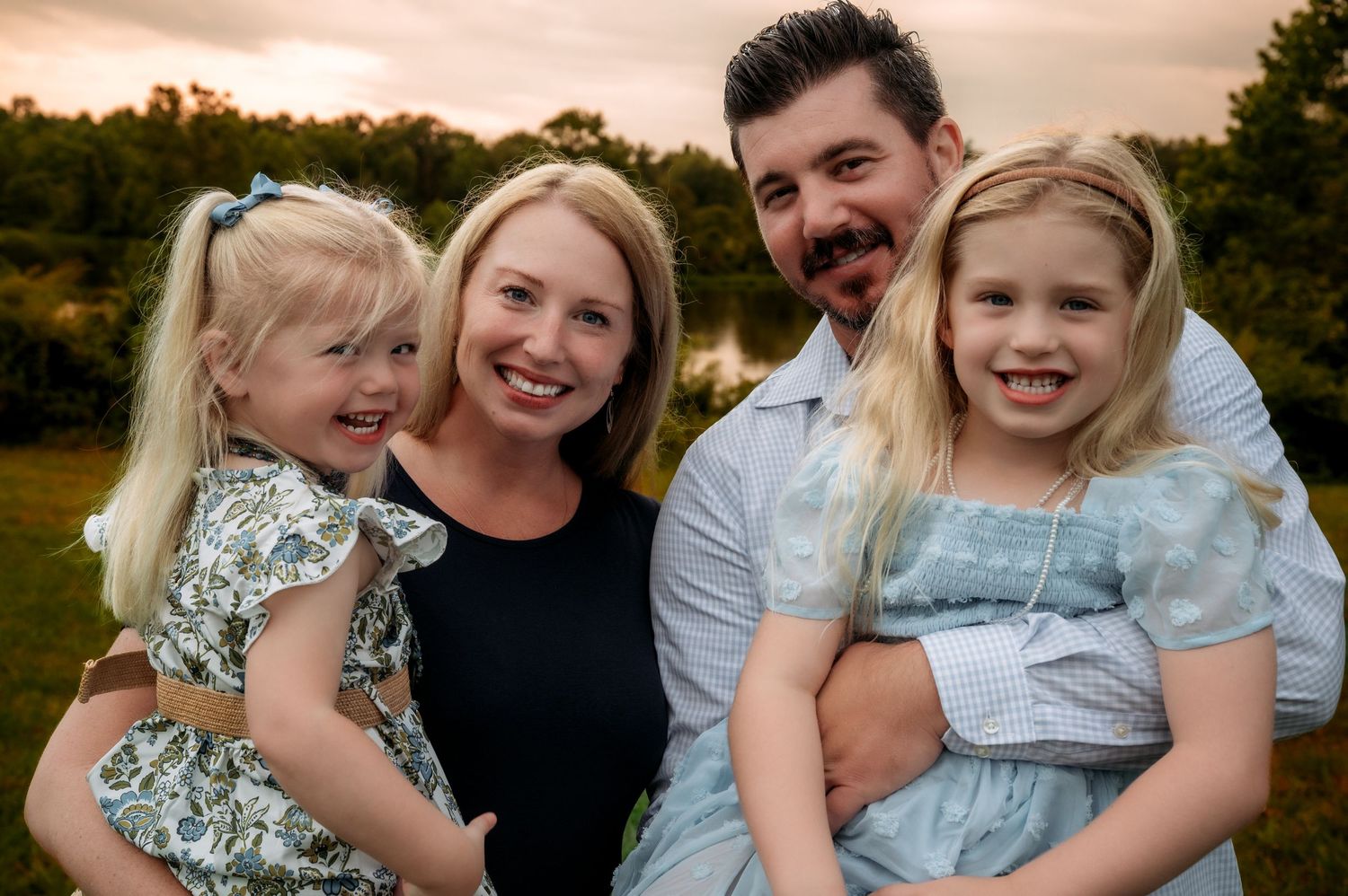 Happy family of four posing outdoors, parents holding two young blonde girls in matching blue dresses at golden hour.