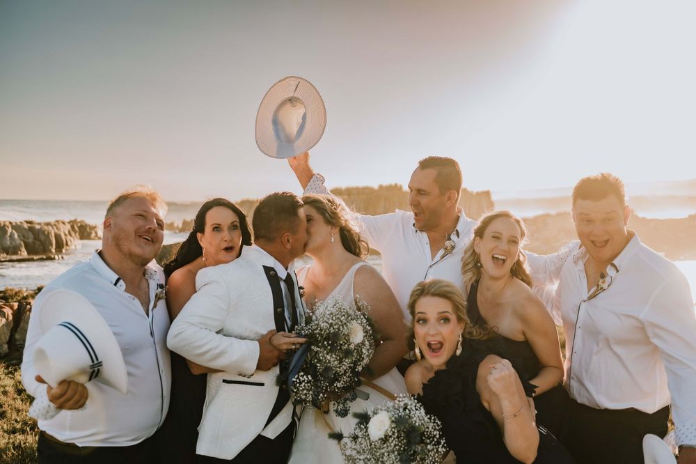 Fun and joyful wedding photo of the bride and groom sharing a kiss while their bridal party celebrates around them at Gourikwa Reserve.