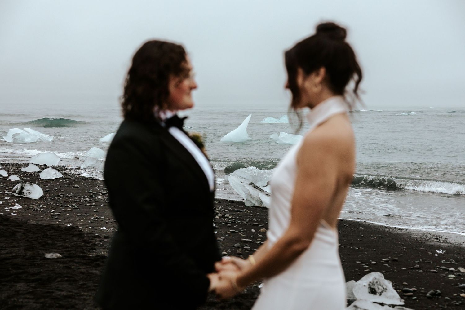 glaciers in focus between two brides while they're blurred out and looking at each other at diamond beach, iceland