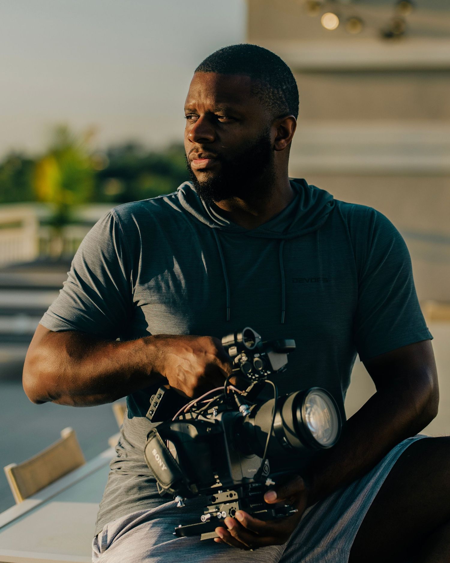 A man holds a professional camera rig outdoors during golden hour, wearing a gray t-shirt on a rooftop.