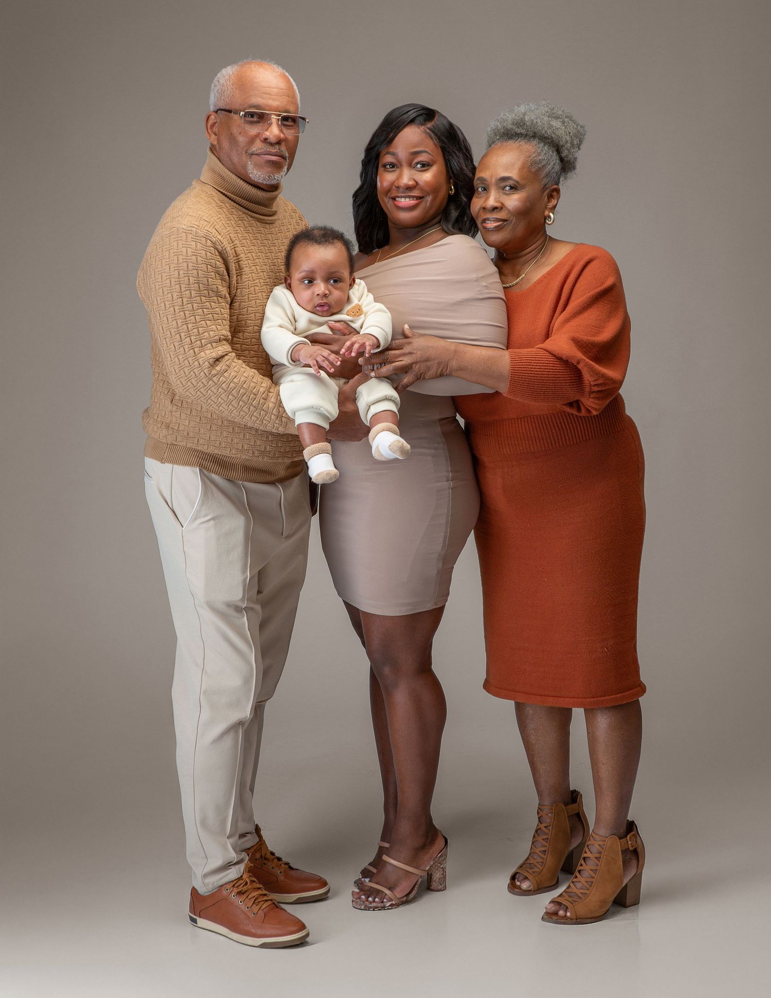 Multi-generational family portrait with grandparents, mother, and baby posing together in warm-toned outfits against gray backdrop.