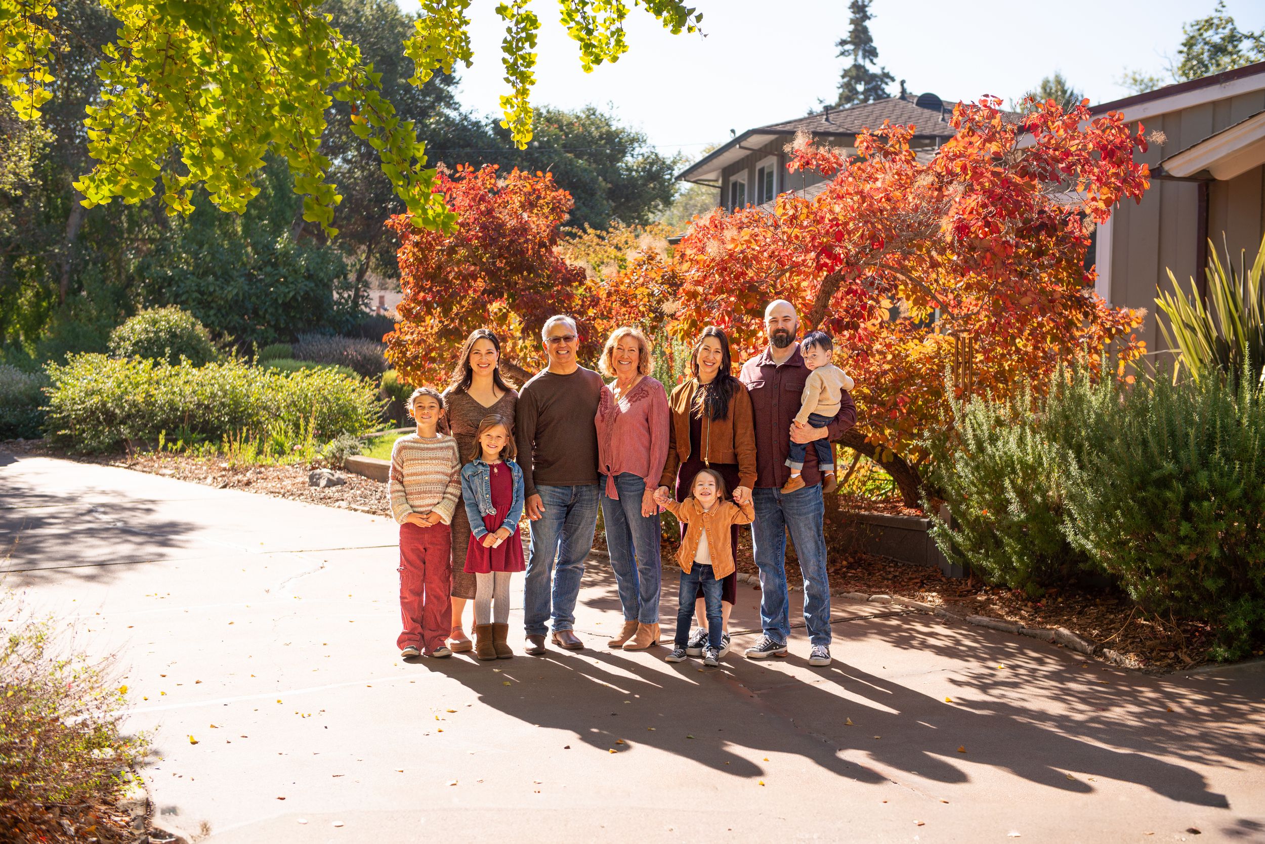 Large family group posing outdoors among vibrant red autumn trees on a sunny day in a garden setting.