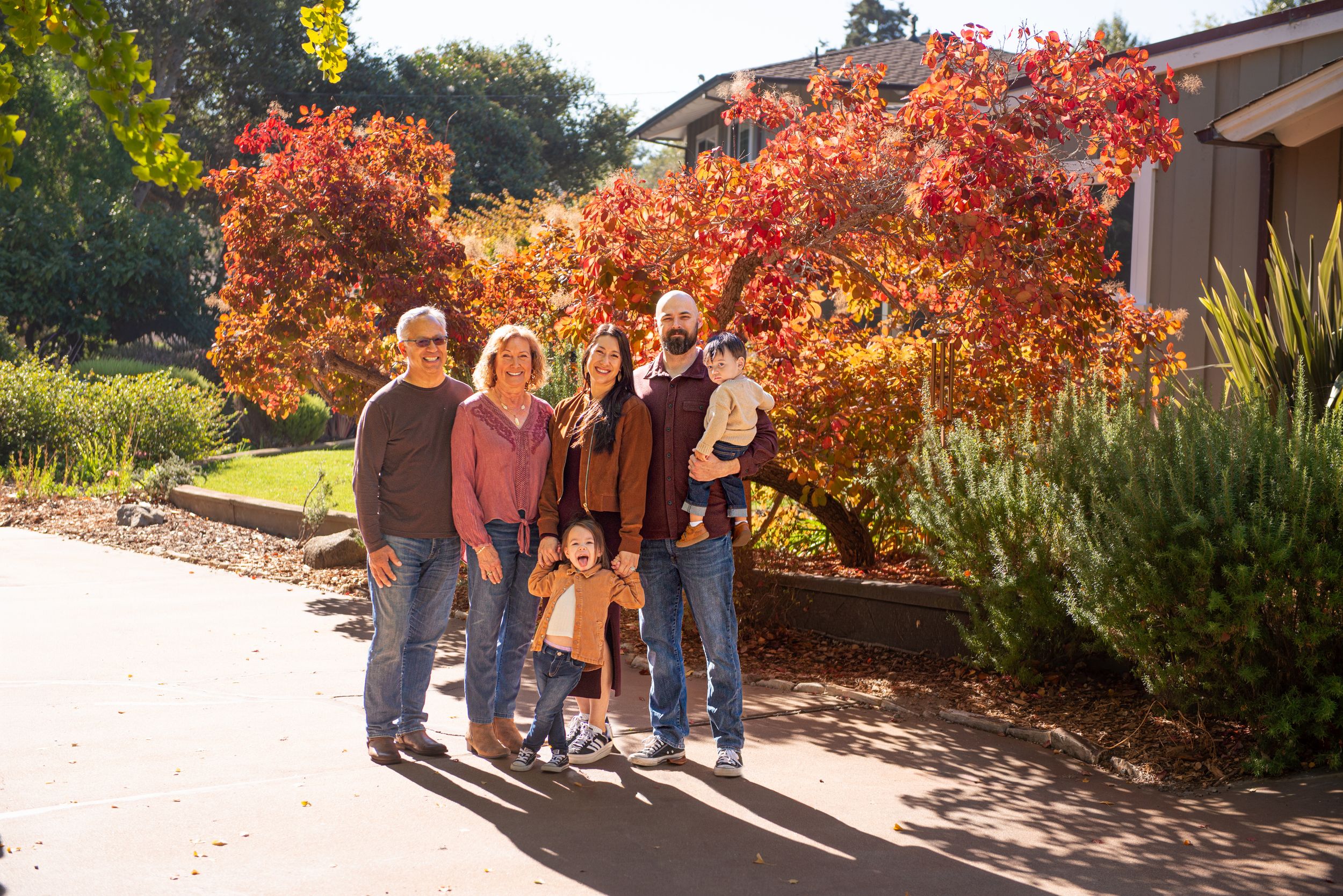 Family of six posing outdoors in front of vibrant red autumn foliage trees on a sunny fall day.