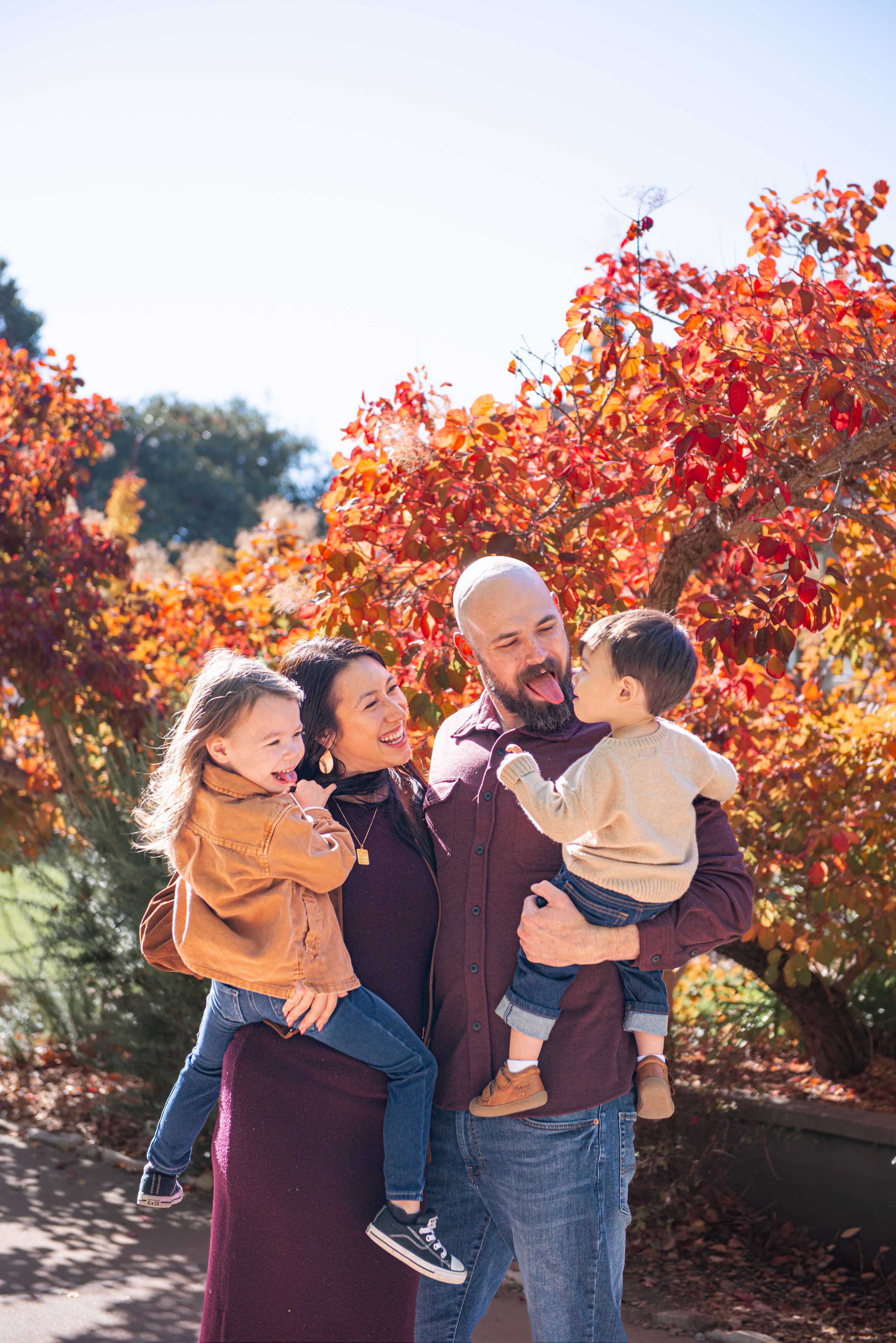 Family of four smiling outdoors in fall, parents holding two toddlers with vibrant red autumn foliage in background.