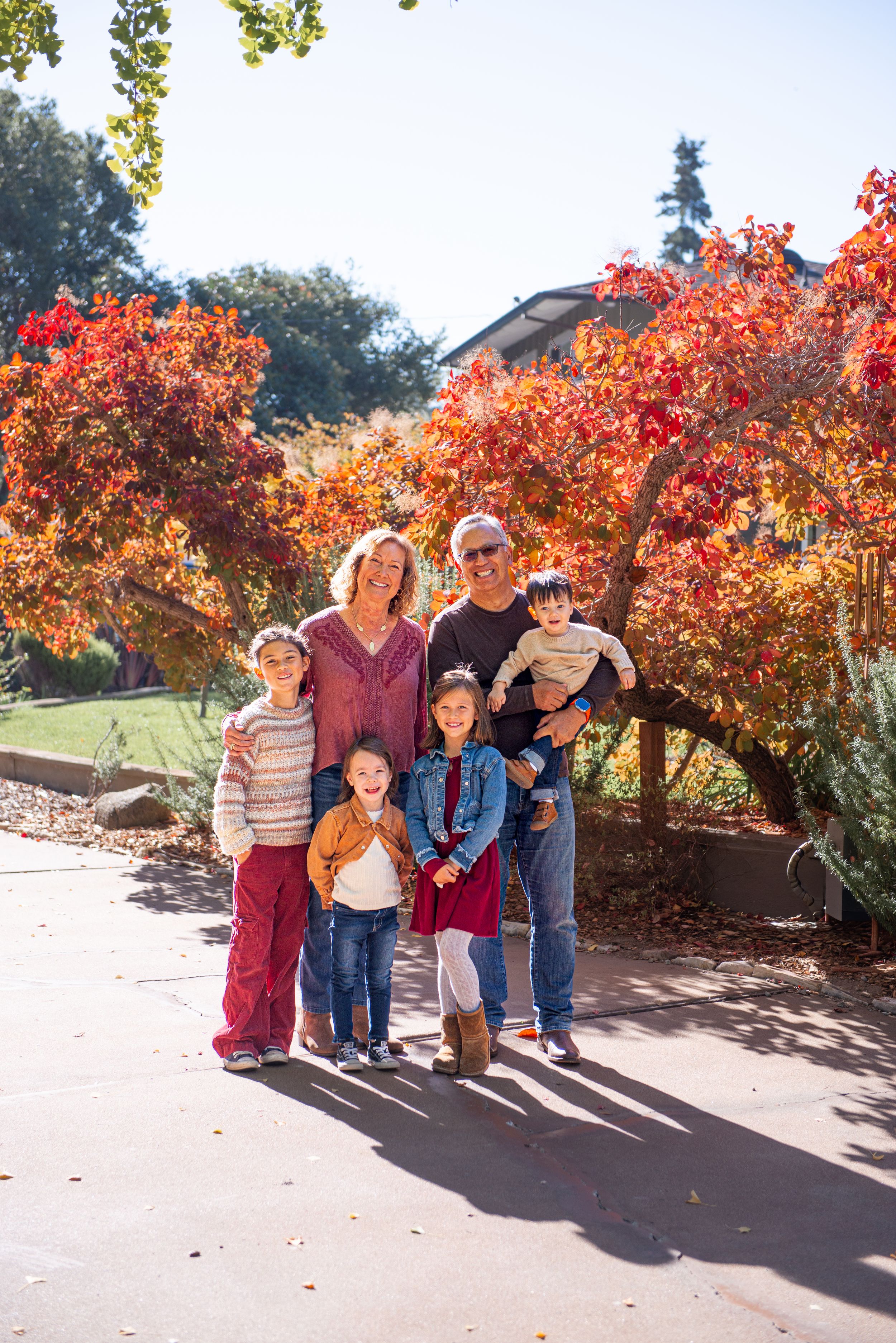 Family of six posing outdoors among vibrant red and orange autumn trees on a sunny fall day.