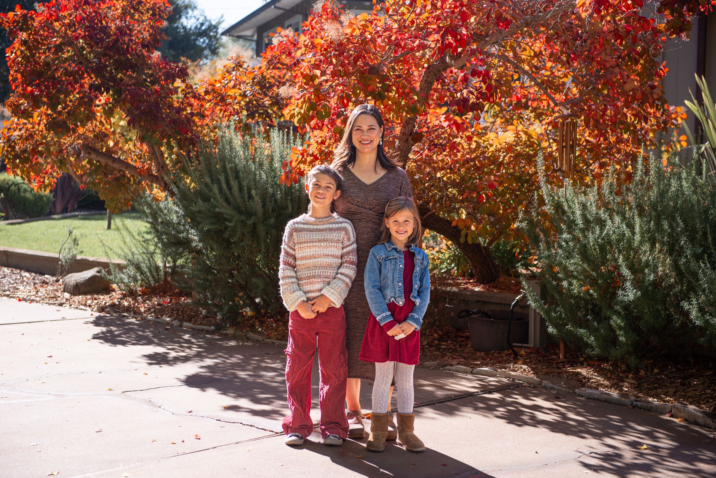 A woman and two children pose outdoors in front of vibrant red and orange fall foliage on a sunny day.