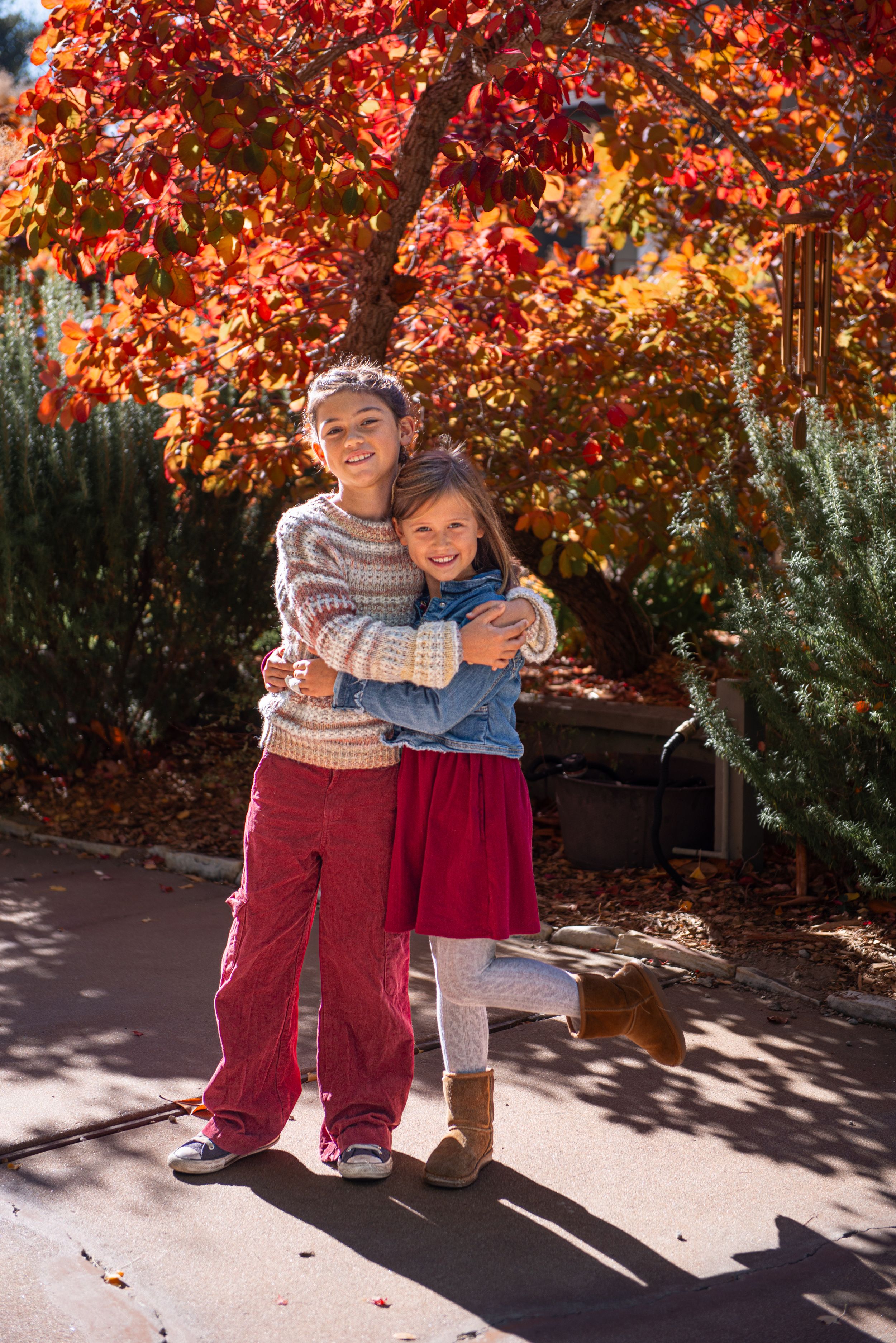 Two girls hugging and smiling outdoors in fall, surrounded by vibrant red autumn foliage on a sunny day.