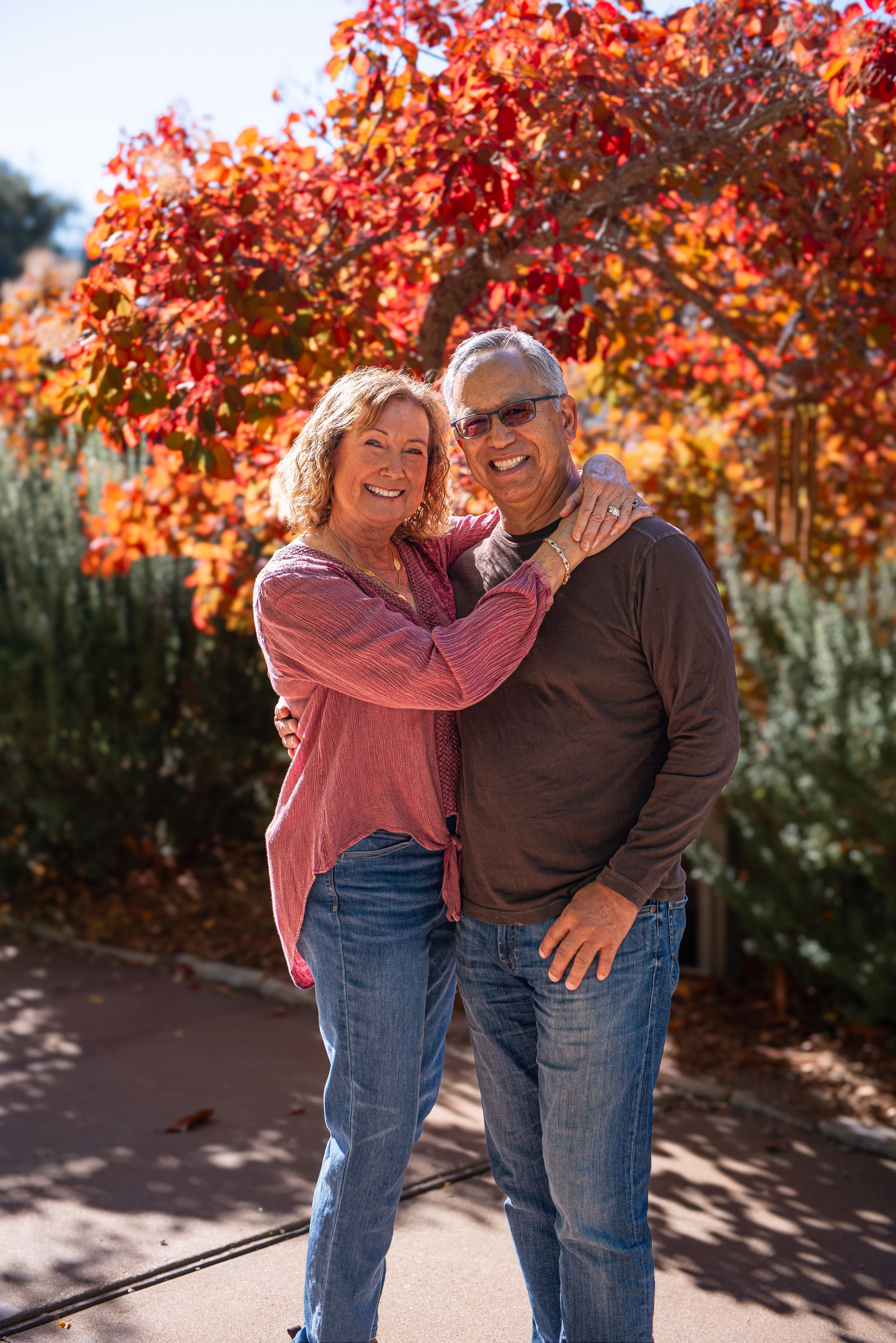 A smiling couple embraces outdoors in front of vibrant red autumn foliage on a sunny fall day.