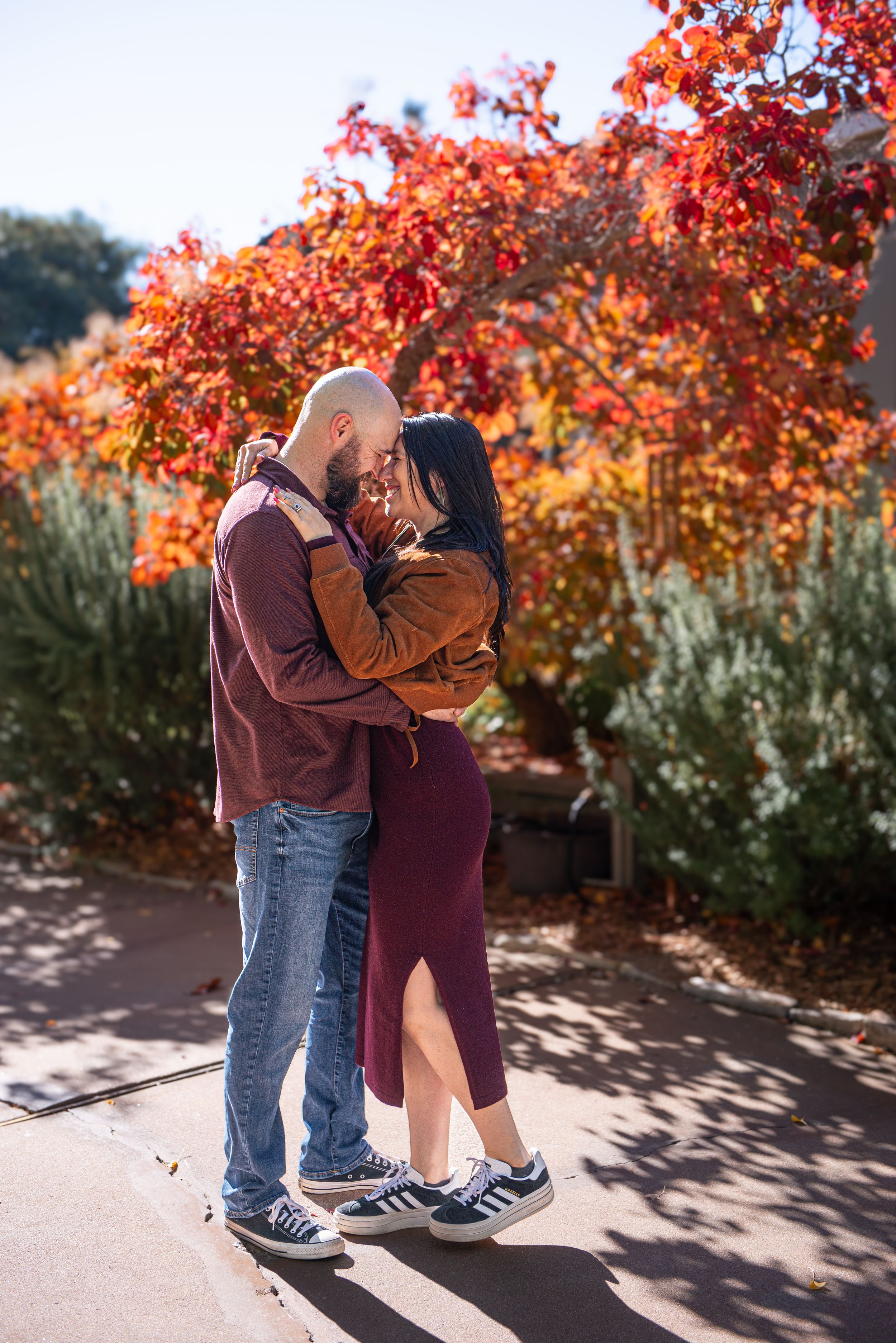 Couple embracing and kissing in front of vibrant red autumn foliage, wearing casual fall outfits outdoors.