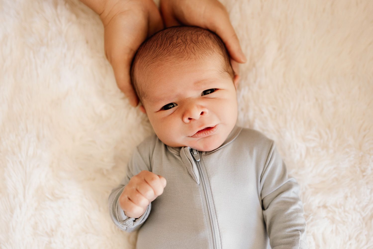 Newborn baby lays on fluffy white blanket with mom's hands gently cradling his head.