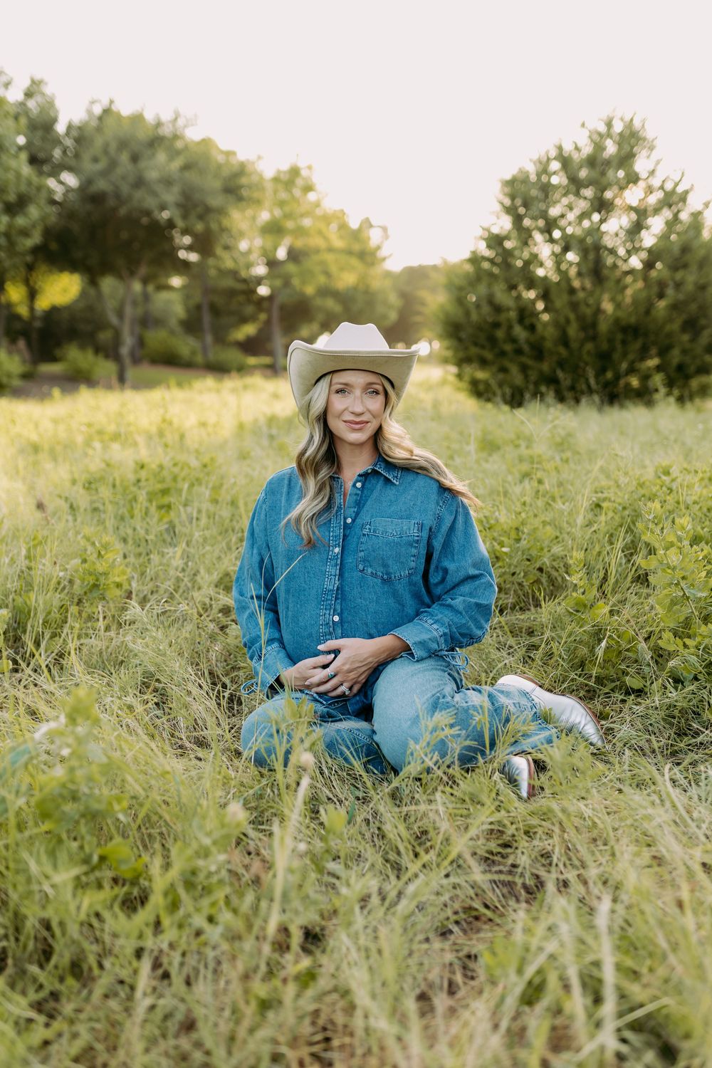 Woman in denim outfit and cowboy hat sitting in green meadow field with trees in background.
