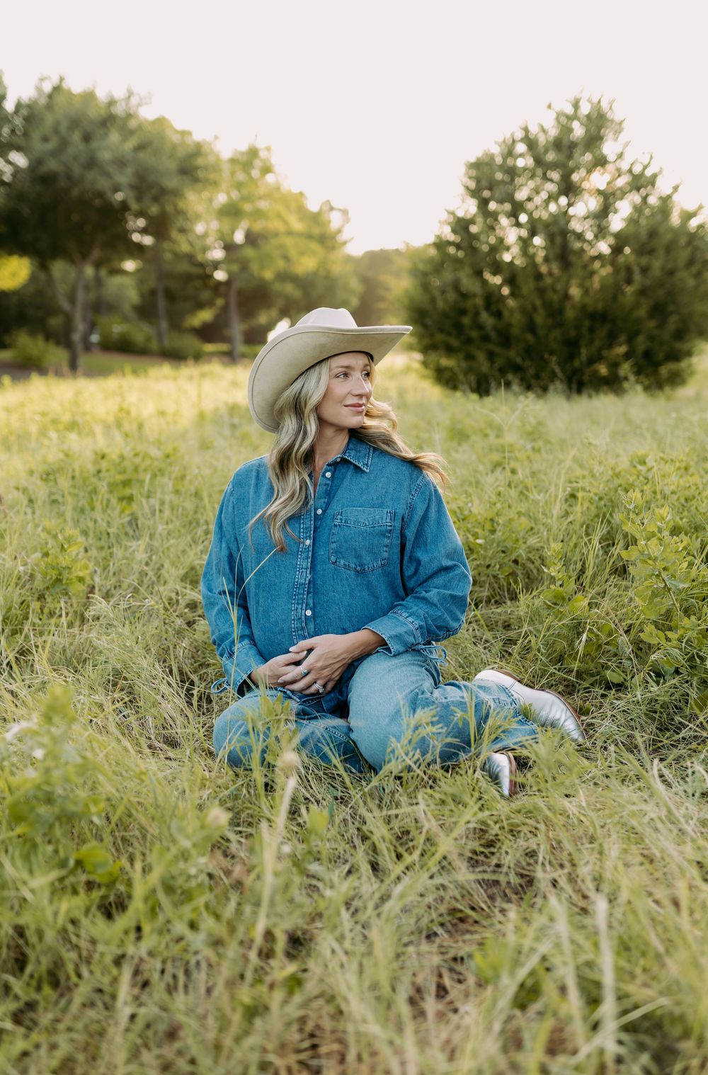 Woman in denim outfit and cowboy hat sitting in green meadow with trees in golden hour light.