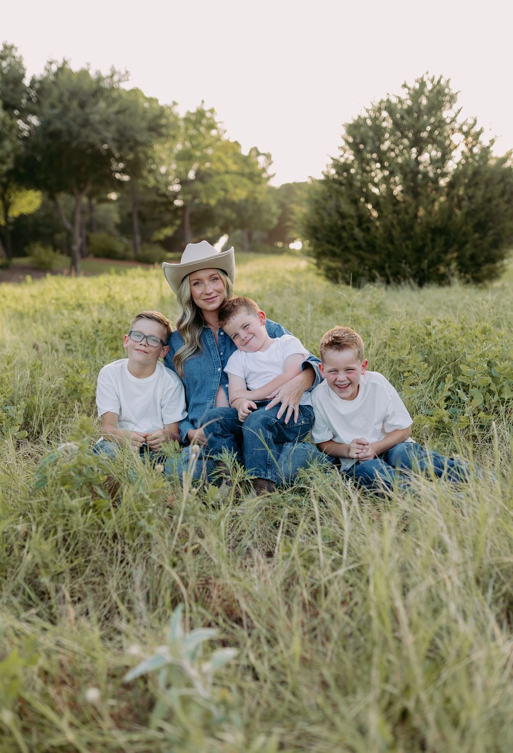A woman in a cowboy hat sits in a green meadow with three young boys in white shirts, smiling for a family photo.