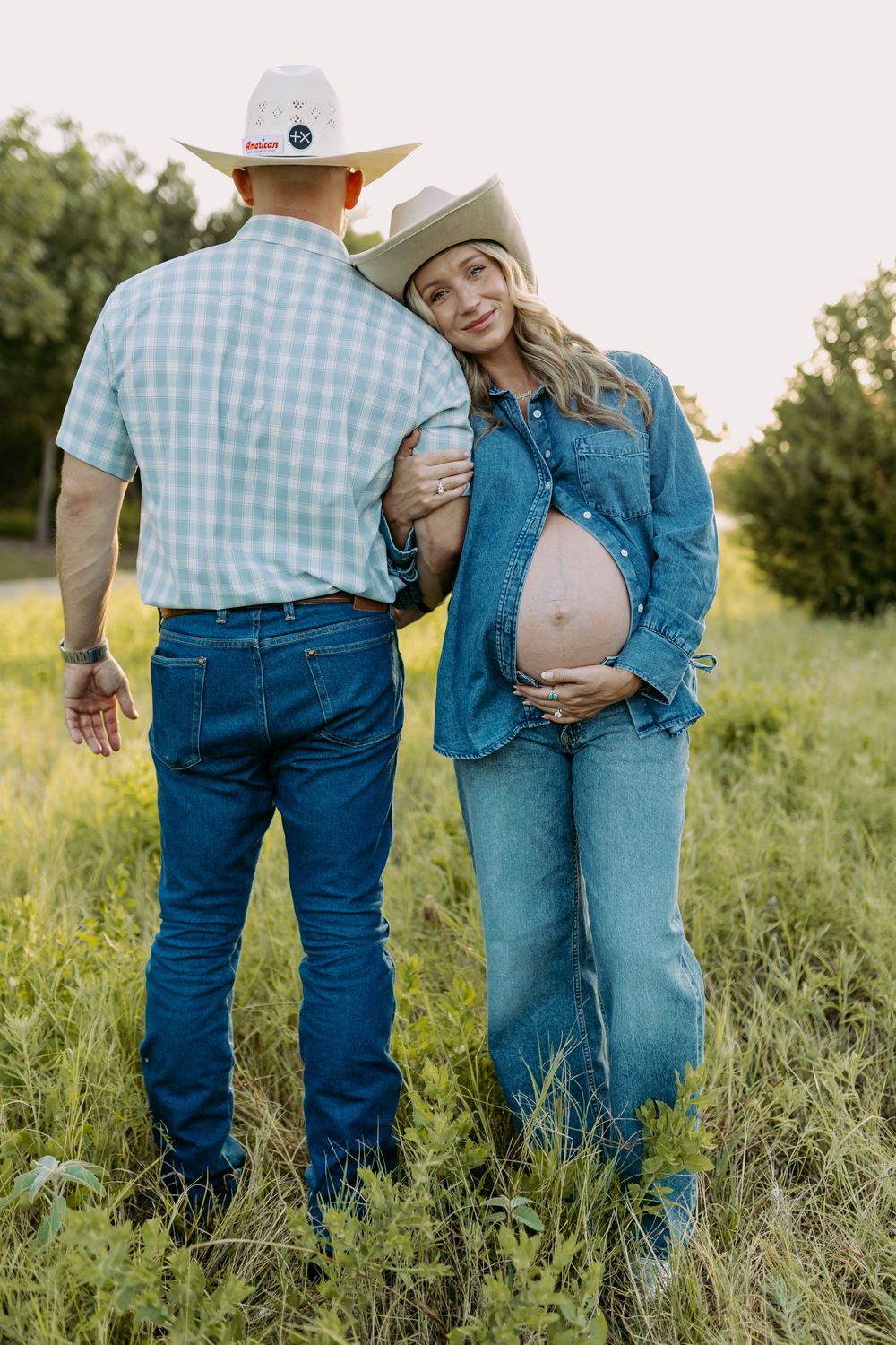 Pregnant couple in cowboy hats and denim standing in a green field, woman cradling baby bump at golden hour.