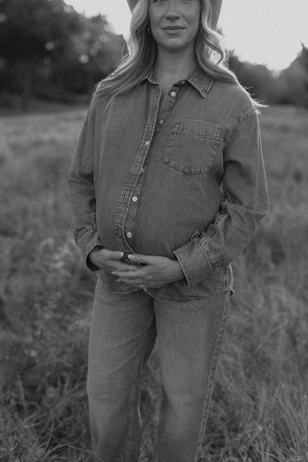 Black and white photo of a pregnant woman in denim shirt and jeans cradling her baby bump in a field.