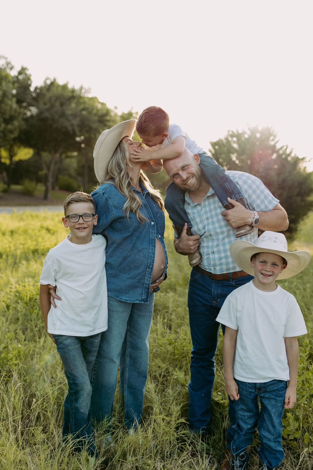 Family of five posing outdoors in a sunny green field, parents and three boys in denim and cowboy hats.