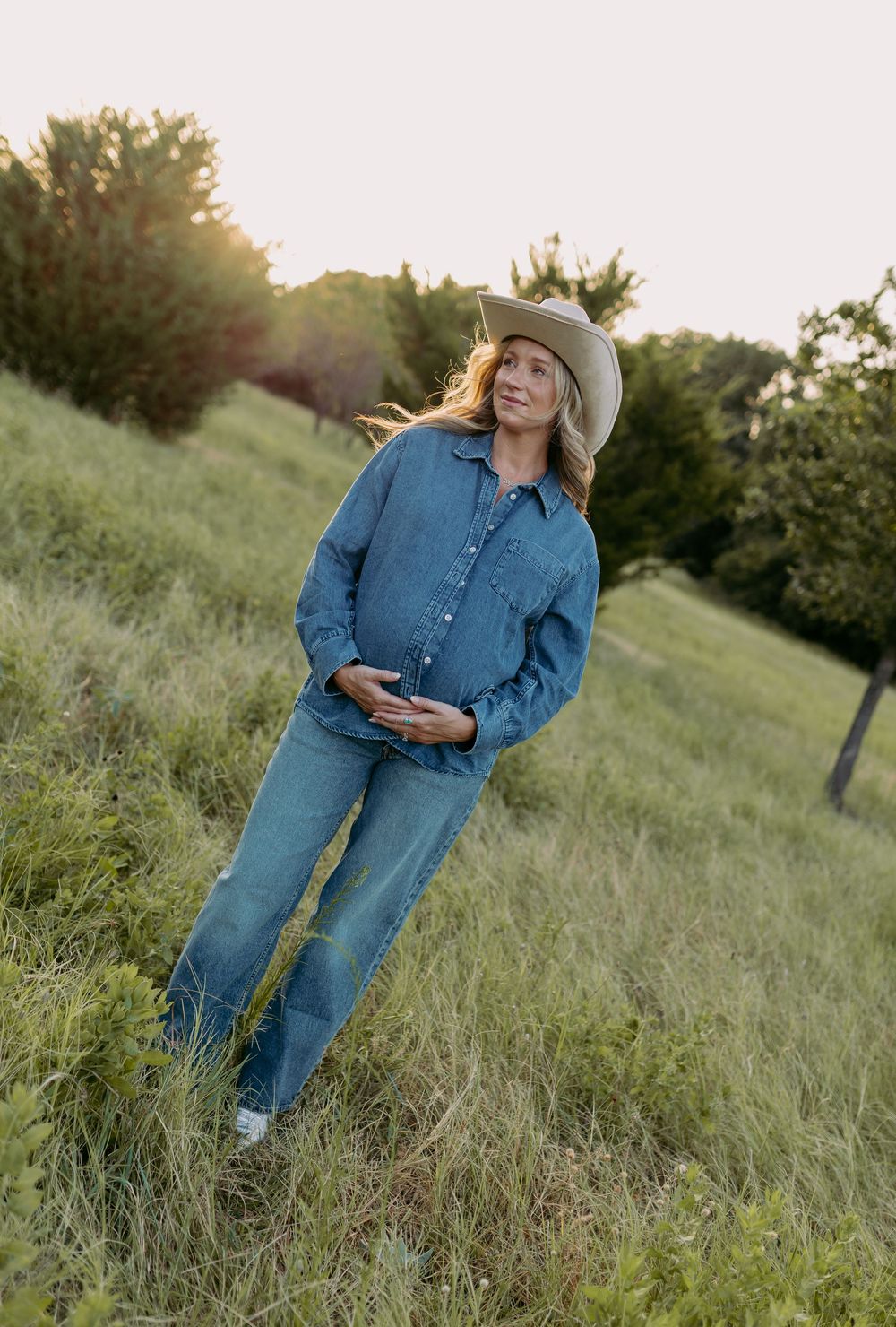 Pregnant woman in denim outfit and cowboy hat standing in green field at golden hour.