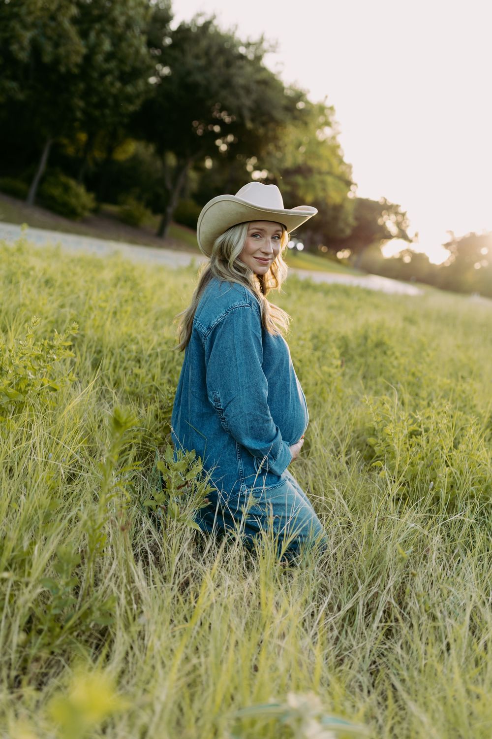 A young blonde woman in a denim jacket and cowboy hat smiles in a lush green field at golden hour.