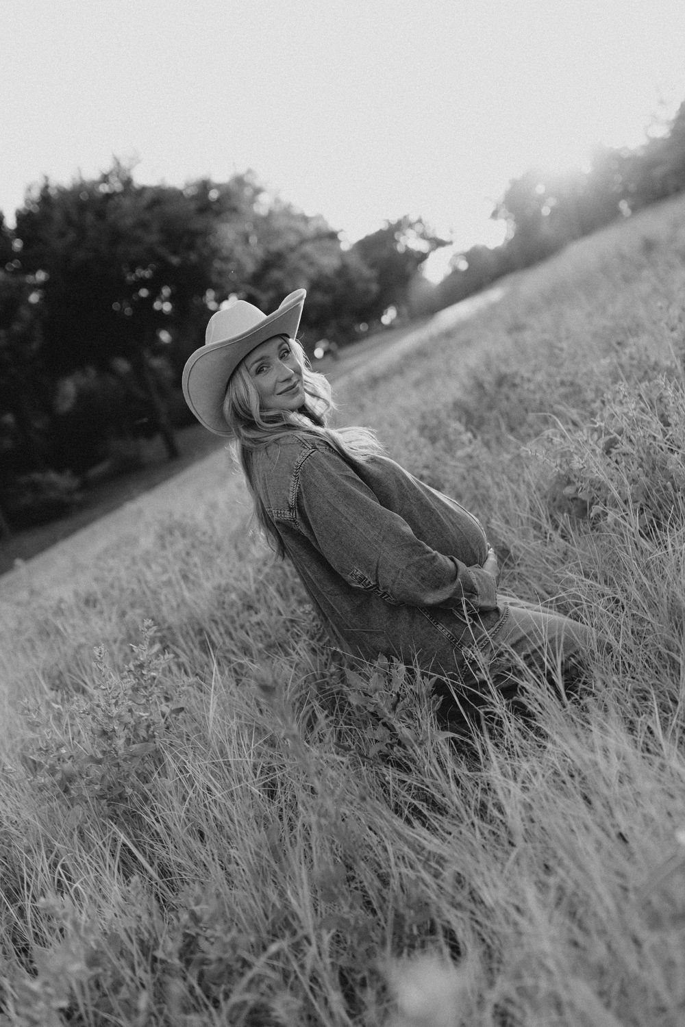 Black and white photo of a smiling woman in a cowboy hat sitting in a grassy field at sunset.