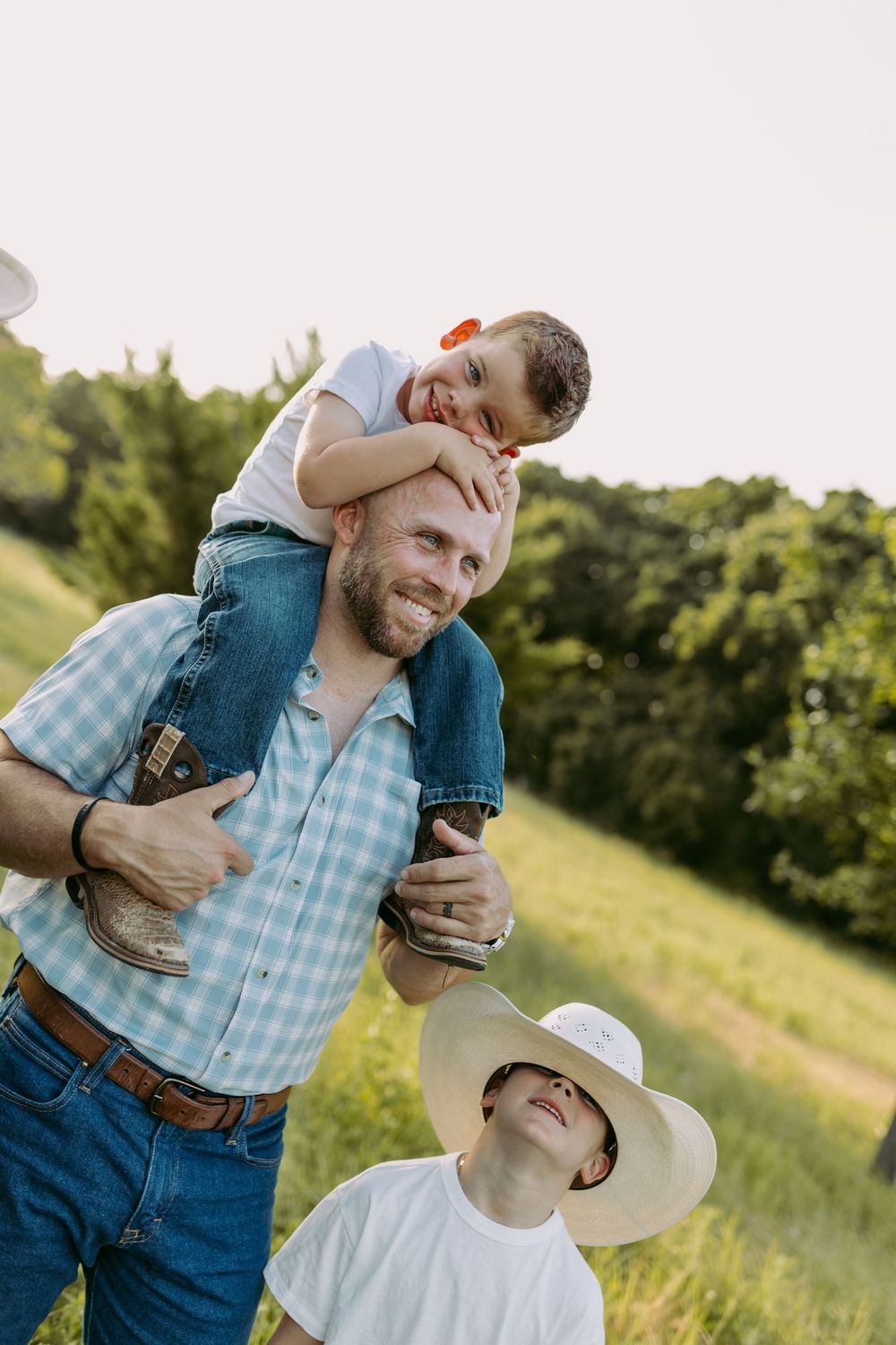 A bearded man carries a laughing child on his shoulders while another child in a cowboy hat looks up in a green field.