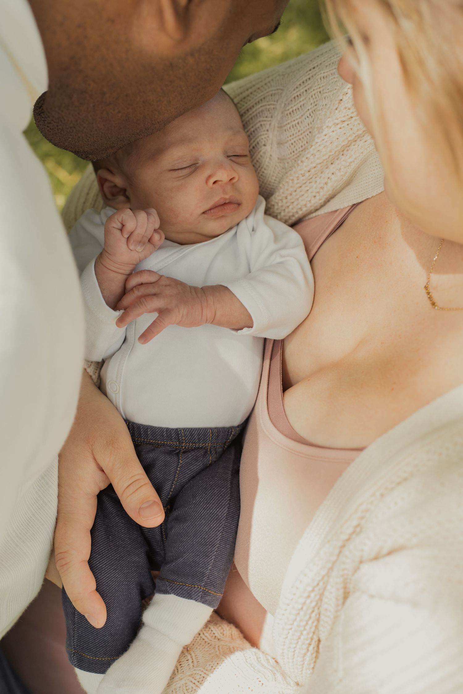 Parents lovingly cradling their sleeping newborn baby outdoors in natural light.