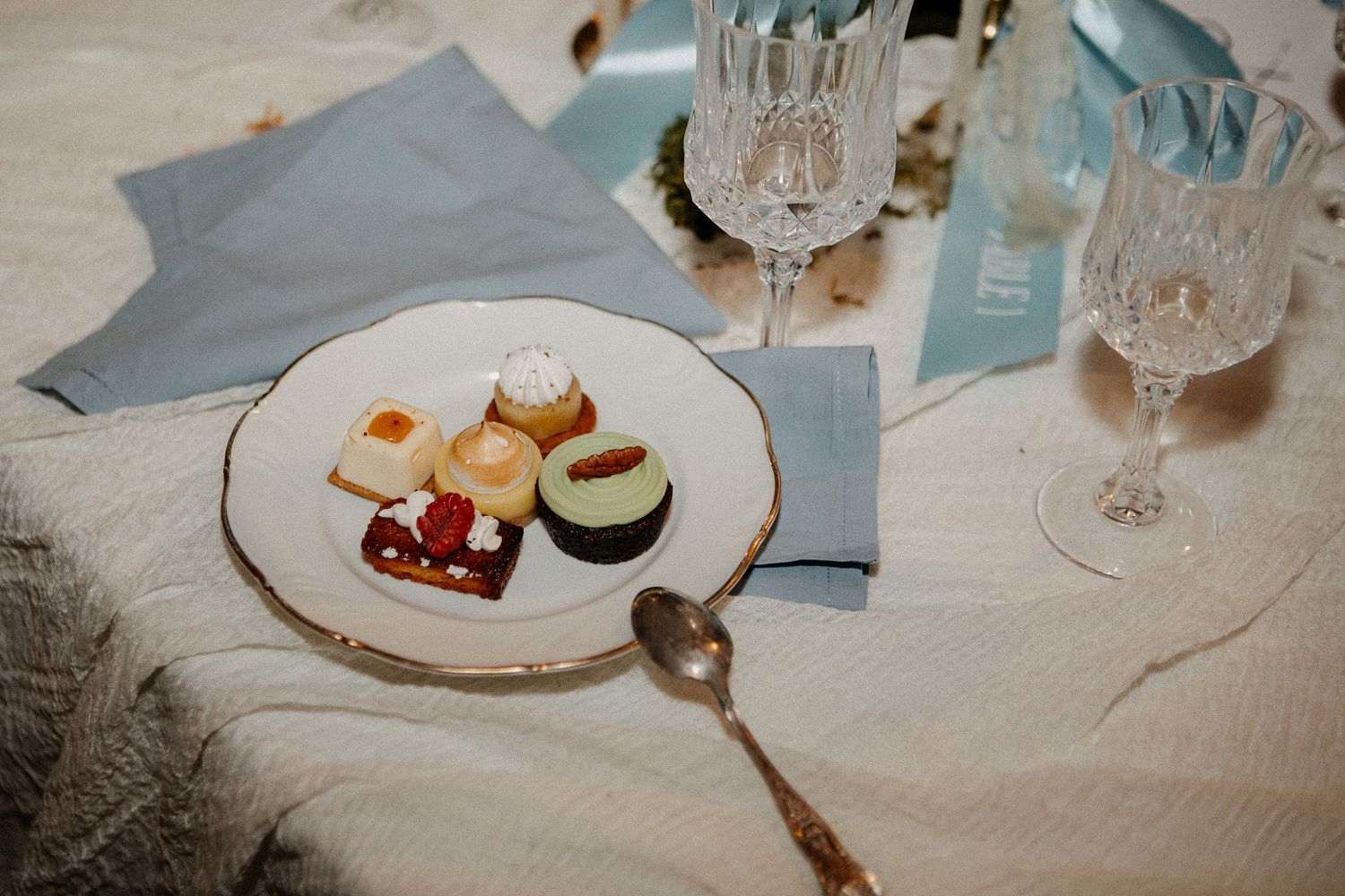 Elegant table setting with assorted petit fours on a gold-rimmed plate, crystal glasses, and light blue napkins.