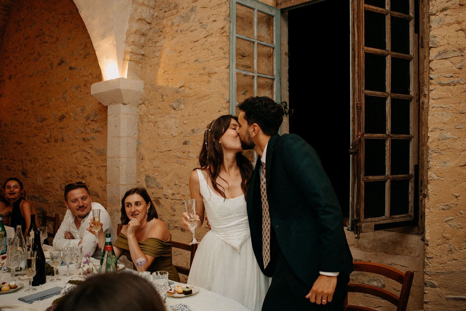 Bride and groom share a kiss at wedding reception dinner in rustic stone venue, guests smiling nearby.
