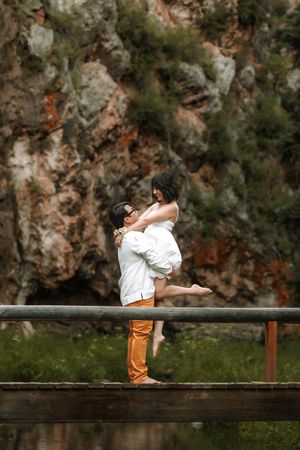 Romantic couple embracing outdoors, man lifting woman near rustic fence with rocky cliff backdrop.