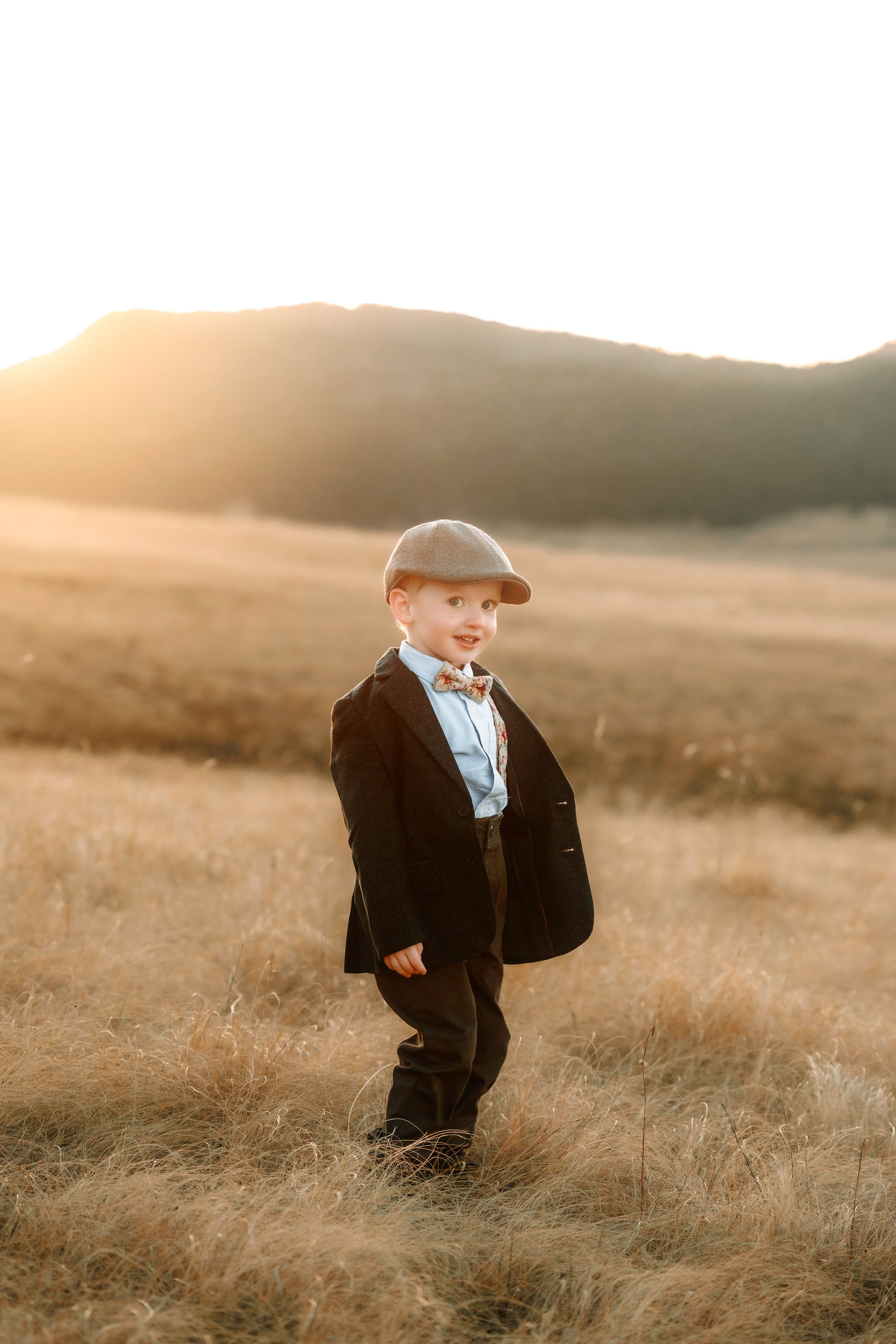 A dapper young boy in a black suit, bow tie, and flat cap stands in a golden field at sunset.