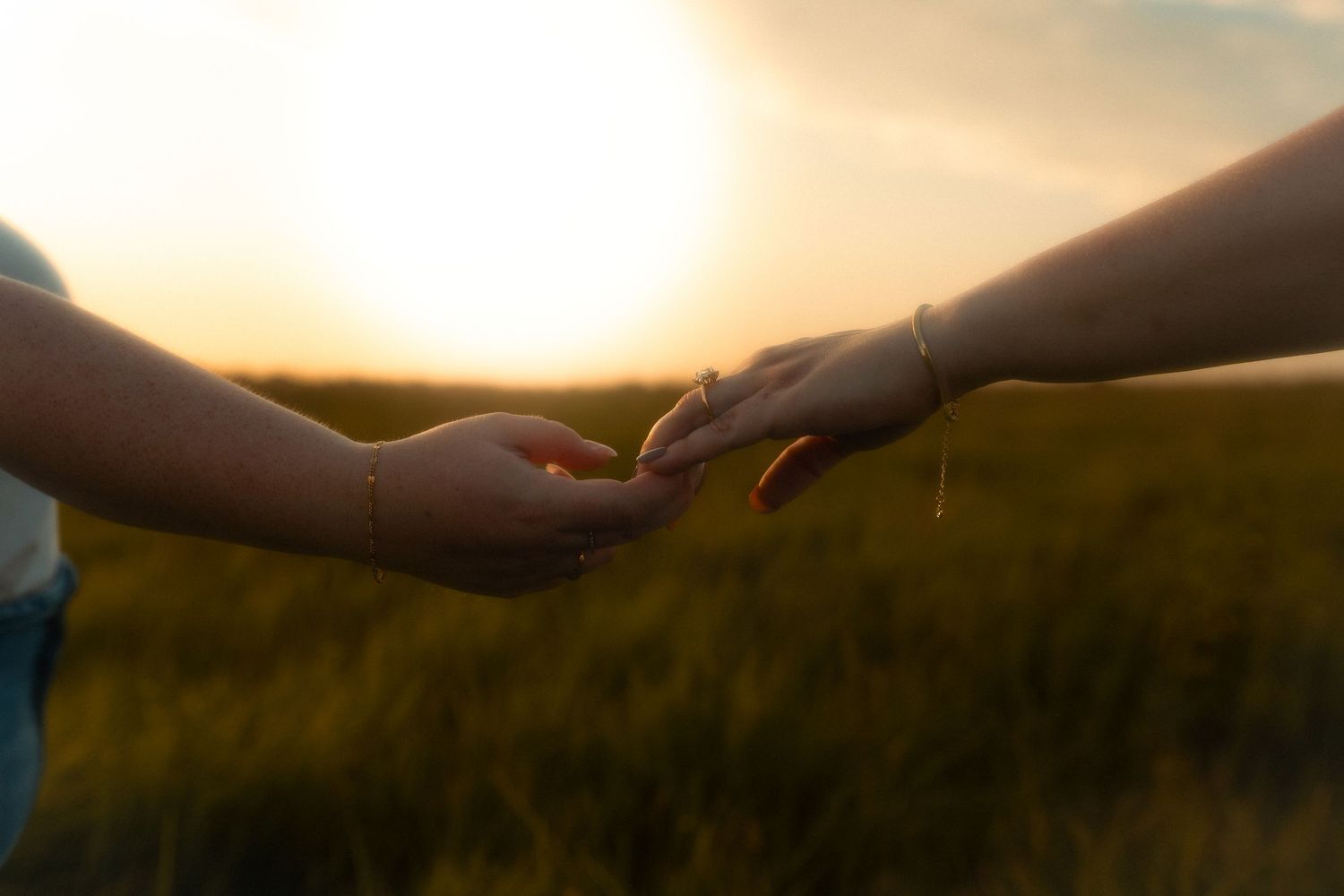 Two hands gently touching at sunset in a green field, symbolizing connection and tenderness in golden light.