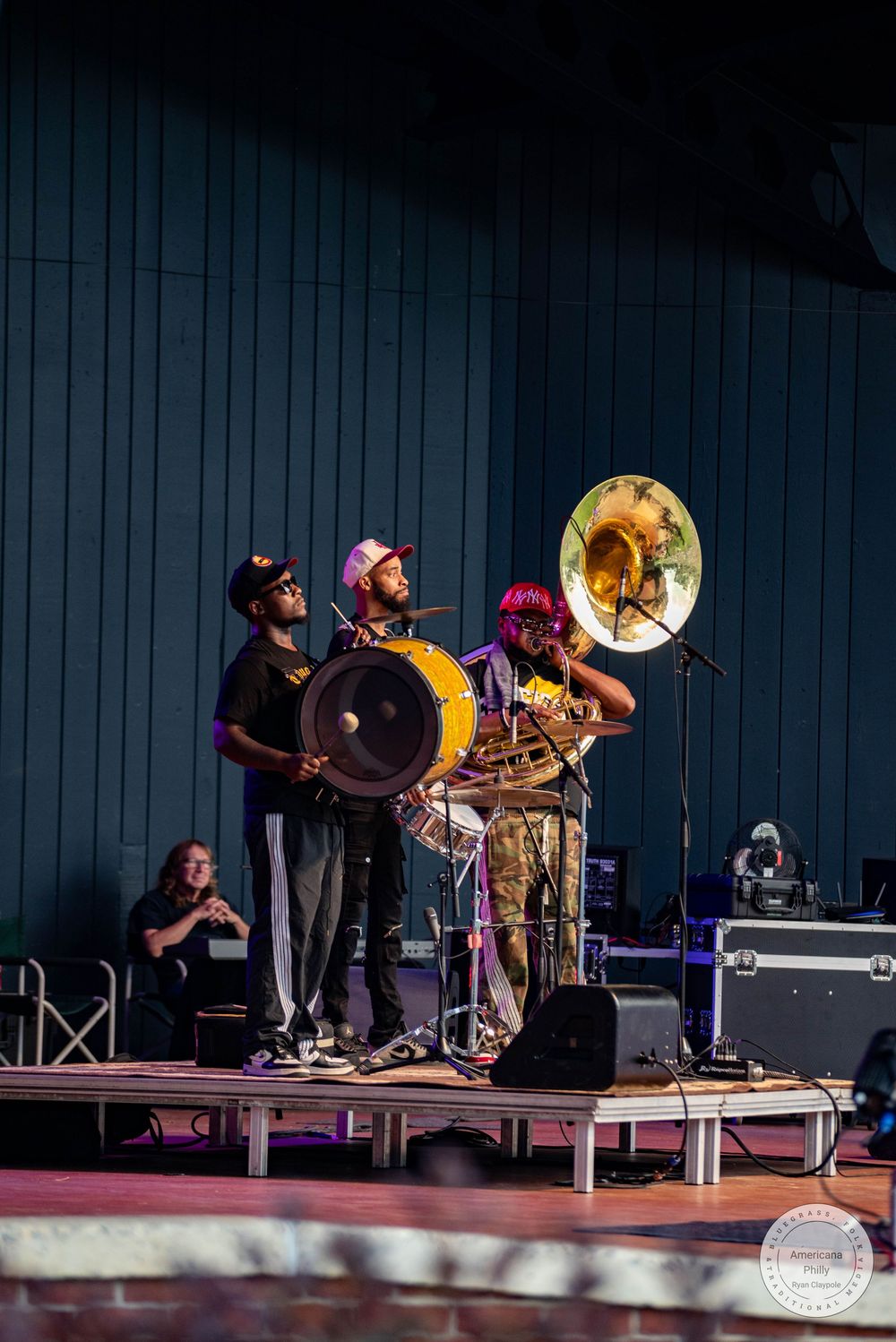 Three musicians perform on stage with a bass drum, trombone, and sousaphone at an outdoor concert.