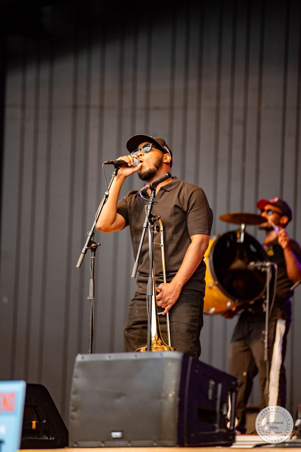 Man in black outfit and cap singing into microphone on stage at outdoor concert.