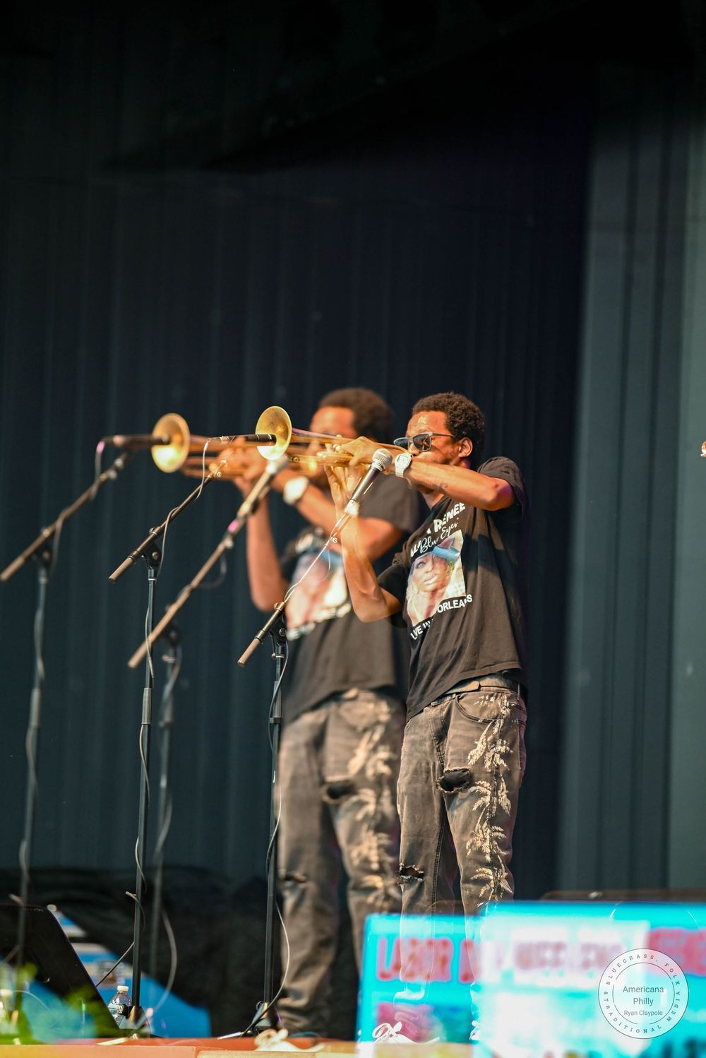 Two musicians playing trombones on stage at an outdoor concert event.