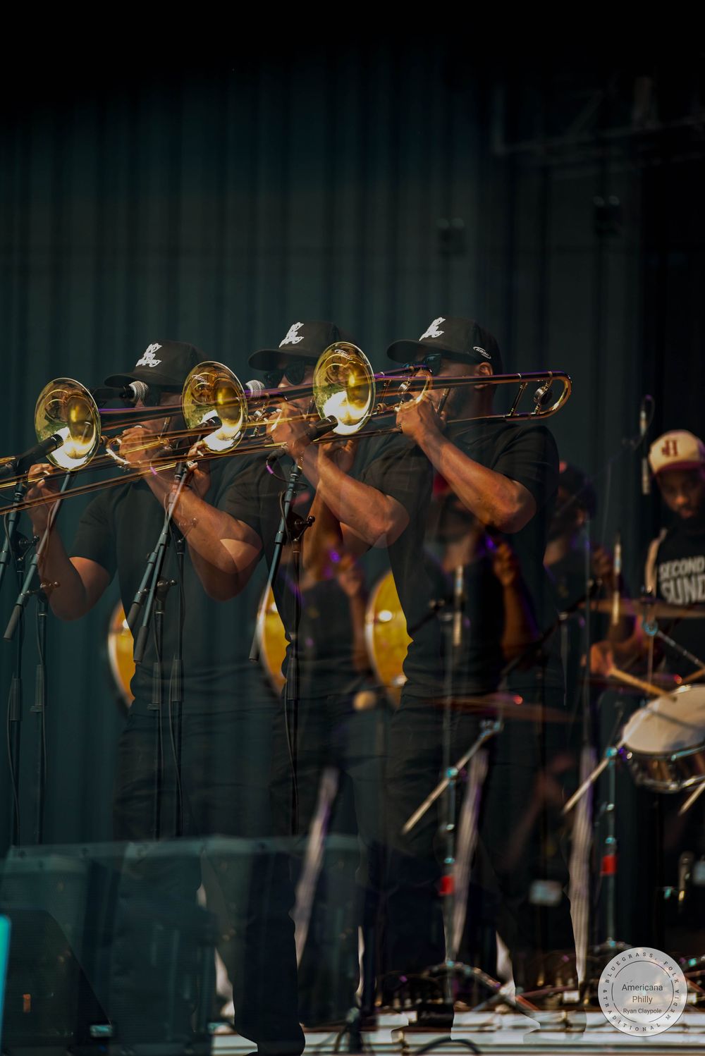 Four musicians in black caps play trombones in unison on stage during a live jazz performance.