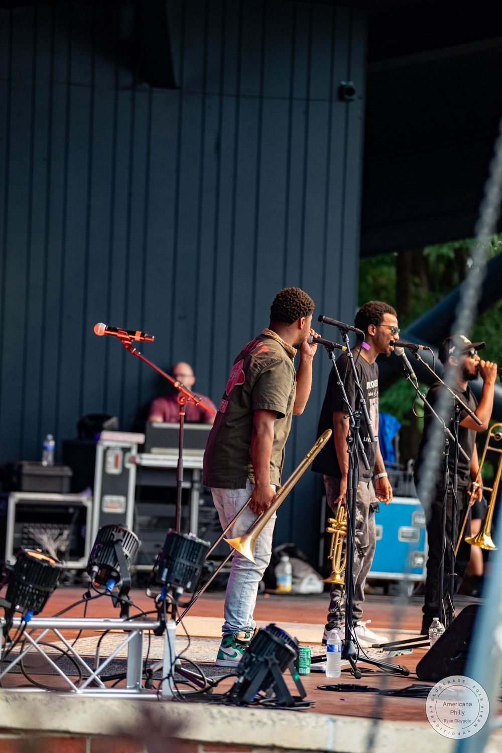Two male singers perform on an outdoor stage with a trumpet player and violinist during a live music concert.