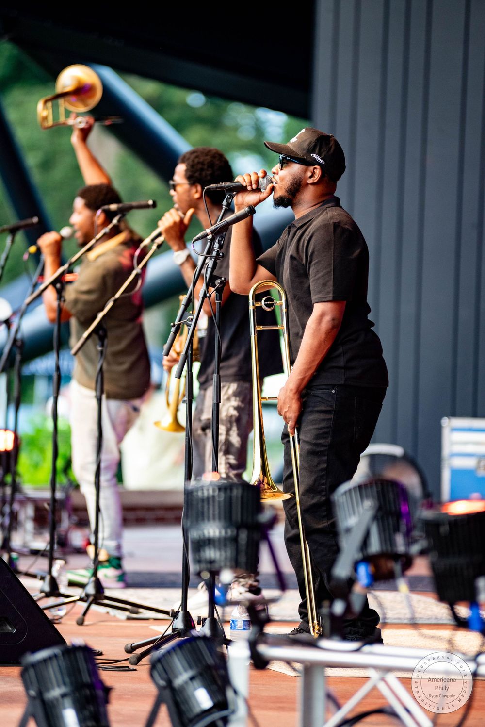 Three musicians perform on stage, two playing trombone and one singing into a microphone at an outdoor concert.