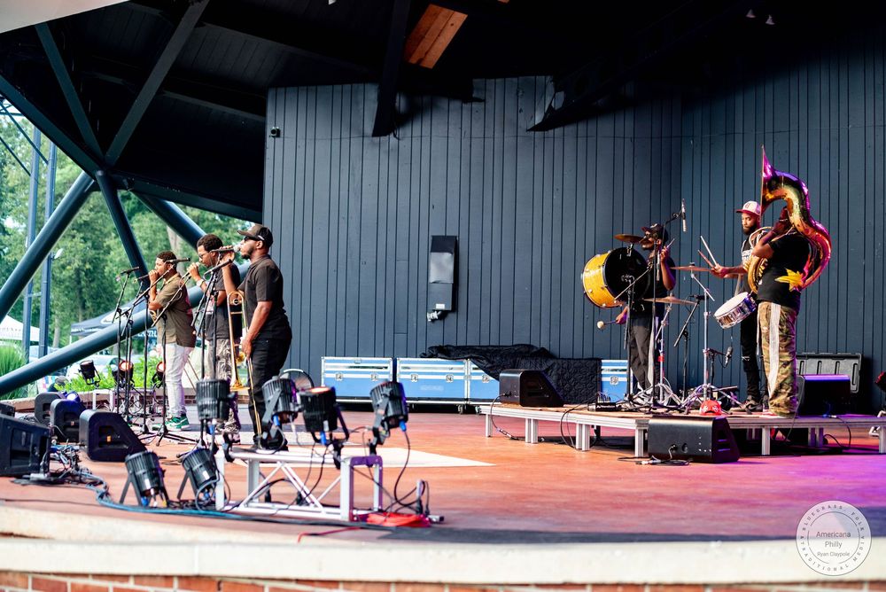 Live band performs on outdoor stage at a park amphitheater on a sunny day, featuring brass instruments and percussion.